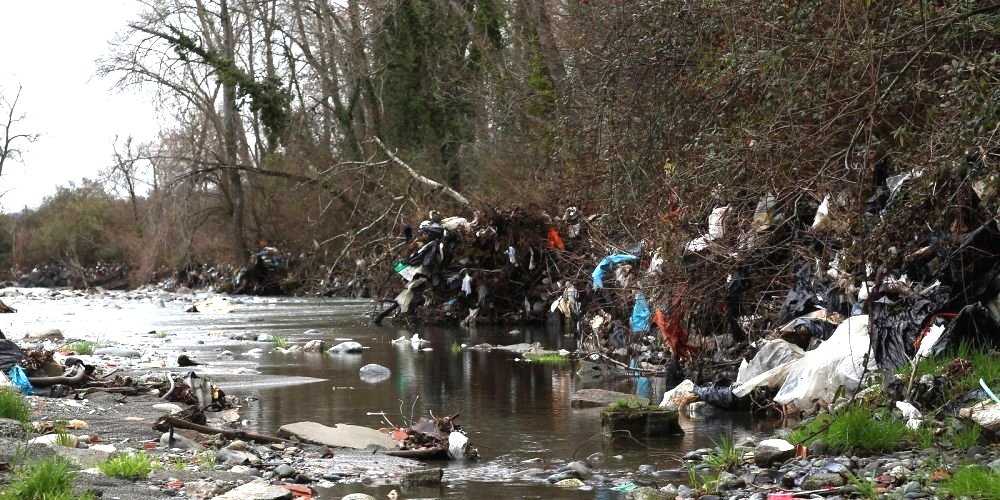 Rivière dont les berges et la végétation sont couvertes de sacs plastique, de bâches et de déchets divers.