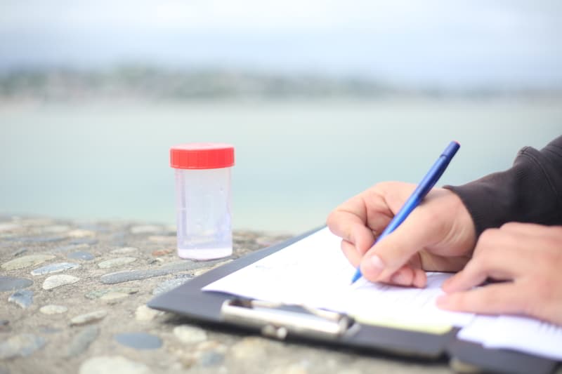 Une personne note des informations sur une fiche à côté d’un flacon de prélèvement d’eau posé sur un muret au bord de la mer
