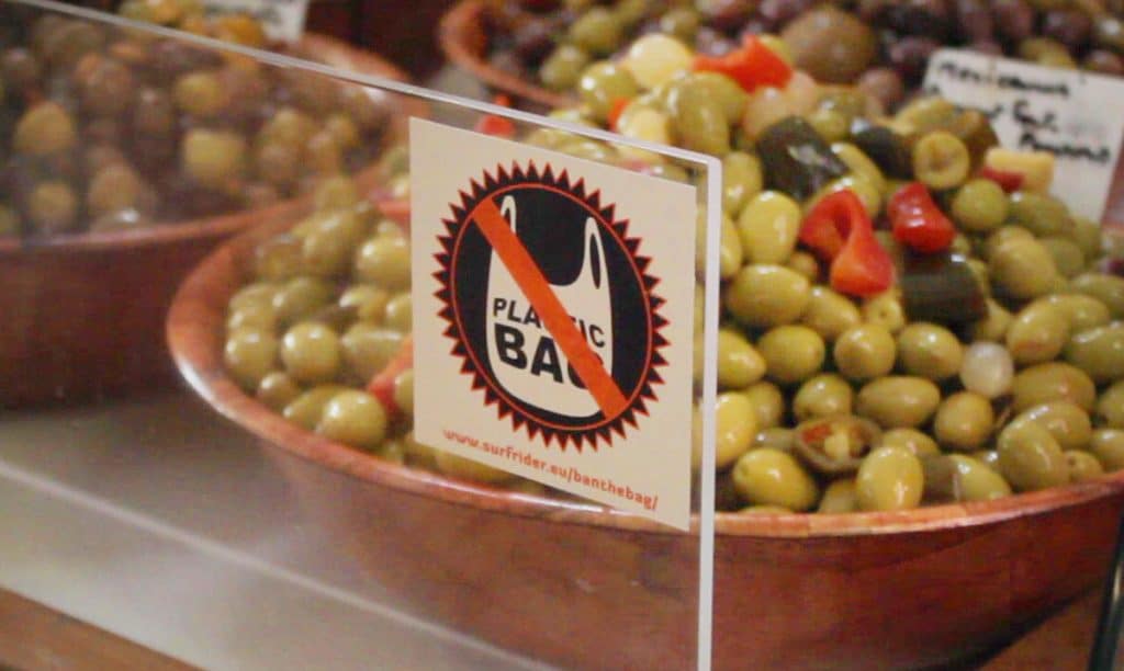 Étal d’olives sur un marché, avec une pancarte « Plastic bag » barrée de la campagne Ban the Bag de Surfrider.