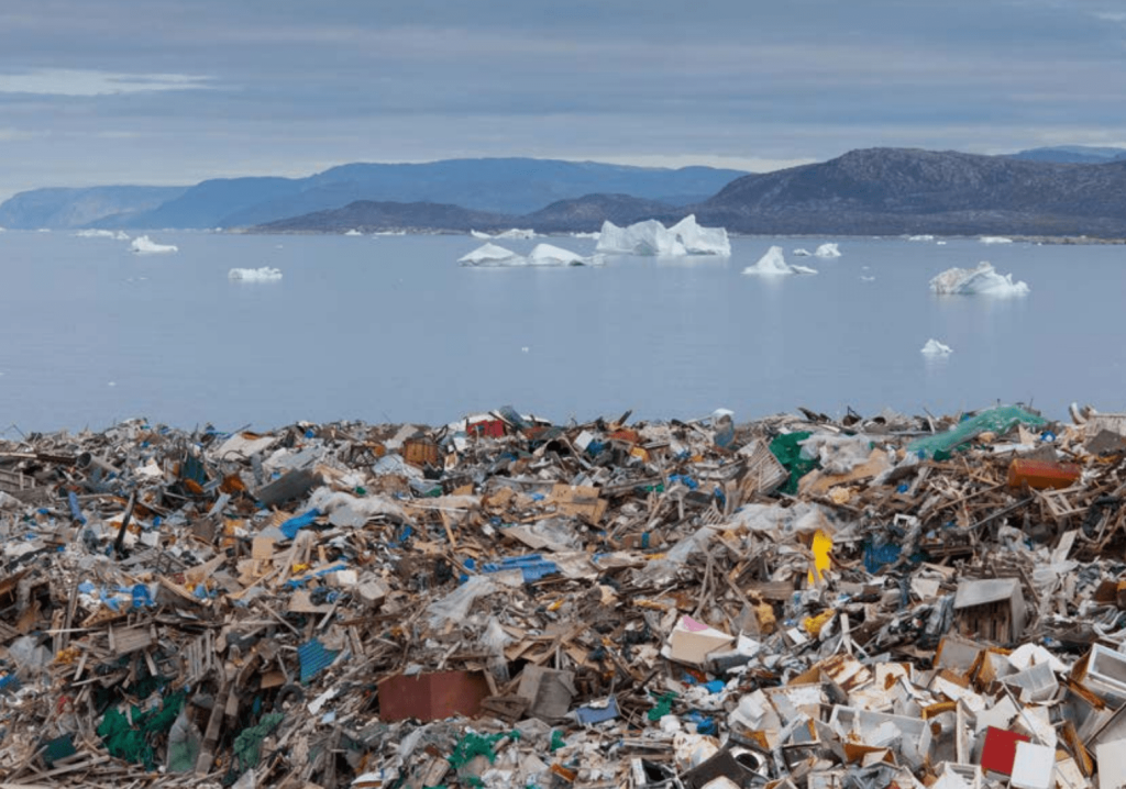 Amoncellement de déchets (plastiques, bois, métal) au premier plan, au bord d’une mer parsemée d’icebergs, avec des montagnes en arrière-plan.