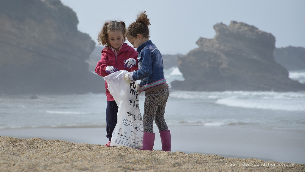 Deux petites filles ramassent des déchets dans un grand sac sur une plage, devant des rochers et des vagues.