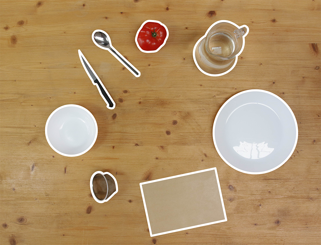 Le matériel sur une table en bois : une tomate, un couteau, une cuillère, un bol, une carafe d’eau, une passoire, une assiette et une feuille de papier.