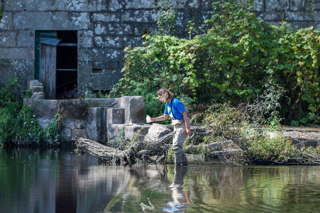 Une bénévole de Surfrider, en tee-shirt bleu et waders, avance dans une rivière au pied d’un vieux moulin en pierre, un flacon de prélèvement à la main.