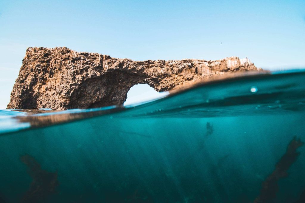 Vue mi-aérienne, mi-sous-marine d’une arche rocheuse au-dessus d’une eau turquoise.