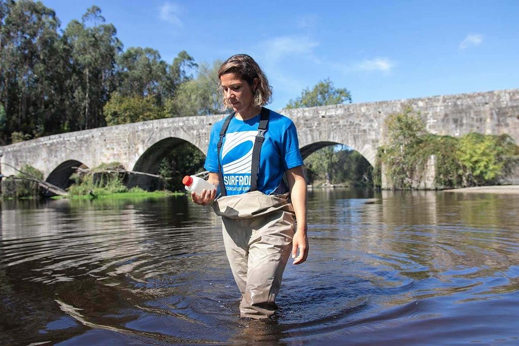 Une bénévole de Surfrider, en waders et tee-shirt bleu, marche dans une rivière en tenant un flacon de prélèvement ; un pont de pierre à arches est visible en arrière-plan.